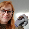 A woman with glasses on smiling while holding up a small bat in her gloved hand