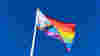 A Progress Pride flag on a flag pole flies in the wind against a bright blue sky.