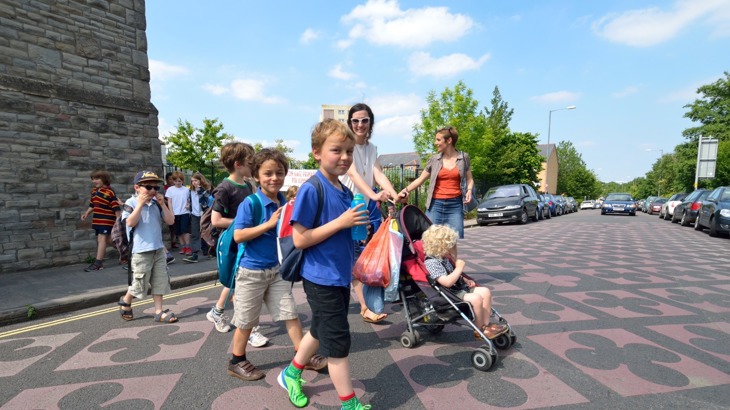 Children walking across a road while a woman pushes a buggy in the background