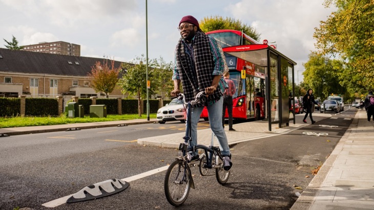 Man in hat and scarf riding fold-up bike in city cycle lane with red double-decker bus in background