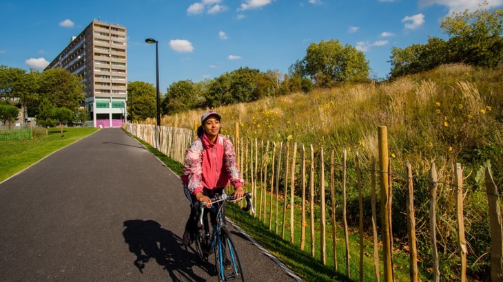A young female cycles along a traffic-free path with a tower block in the background 