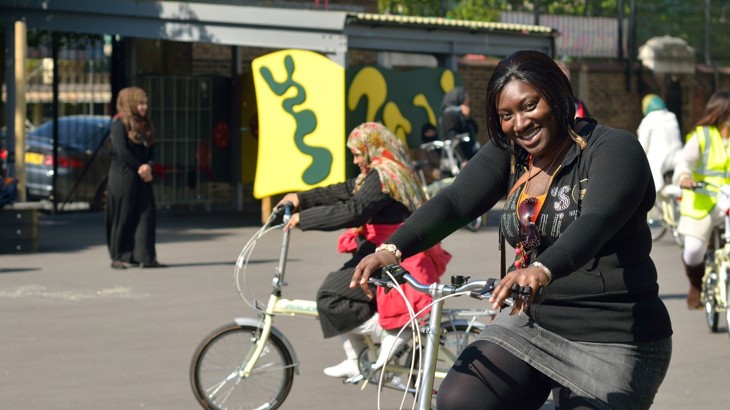 Woman with bicycle at a bike ride training course in a school playground with other adults cycling in background