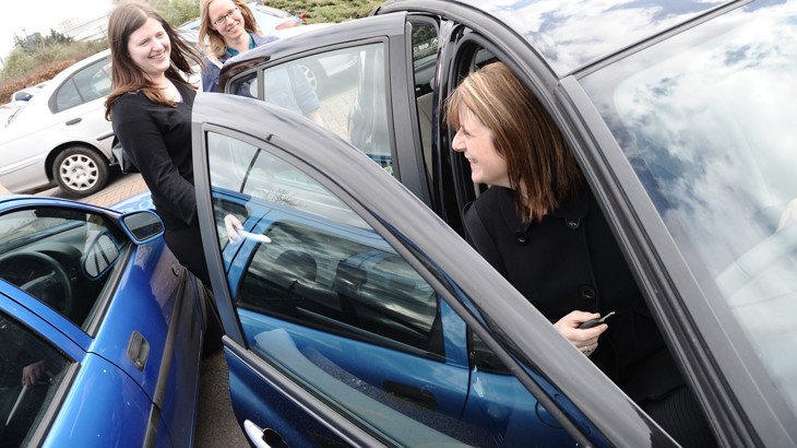 Three women chat outside of a car in a car park 
