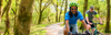 Two men cycle on a lush woodland trail smiling.