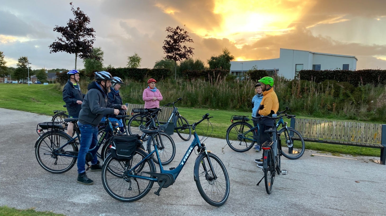 Adults wearing helmets stop on their bikes to listen to instruction from the Walk Wheel Cycle Trust ride leader with the sun setting behind the traffic-free path.