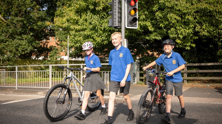Children in school uniform walking and pushing bikes across a toucan crossing on a sunny day
