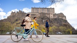 Three people walking, one with a bike, on the pavement with Edinburgh castle on the hill in the background.