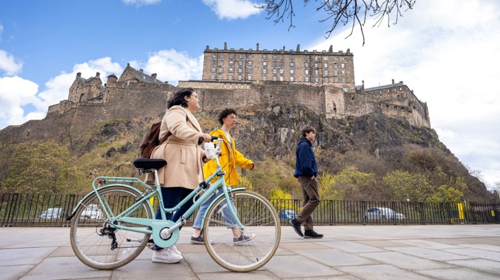 Three people walking, one with a bike, on the pavement with Edinburgh castle on the hill in the background.