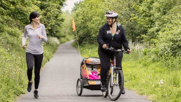 A runner and a cyclist pulling a pink trailer with child in it on traffic-free greenway