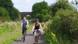 Man and woman cycling on greenway with bridge in the background