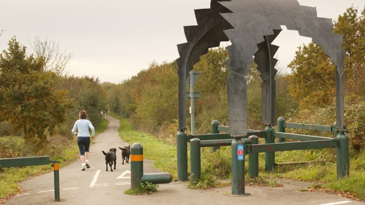 Runner with two dogs on tarmac path through woods with gazebo sculpture to the right side