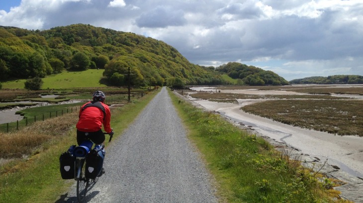 Cyclist in red jacket on gravel path by estuary with green hills and trees to the side