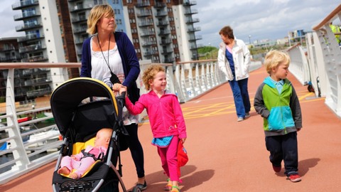 Woman with baby in pushchair with two small children and older woman, walking over bridge in urban area