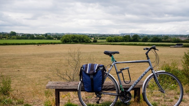A road bike with a blue saddle bag is leant up against a backless wooden bench. Behind is a flat agricultural field on a sunny day in the countryside.