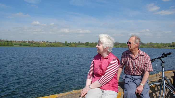 Older man and woman sitting on stone wall by reservoir with bike leaning