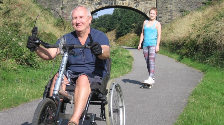 A man on a hand tricycle and girl on a skateboard on tarmac greenway