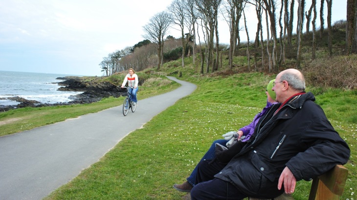 Couple sitting on park bench, looking at cyclist on coastal cycle path
