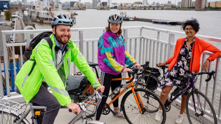 Three cyclists in brightly-coloured jackets, two of them wearing helmets, by the Thames in London