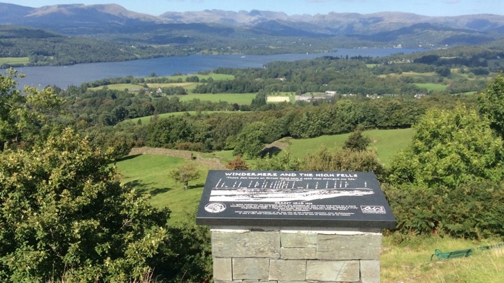 View of Lake Windermere with hills and trees in background and interpretation board in the foreground