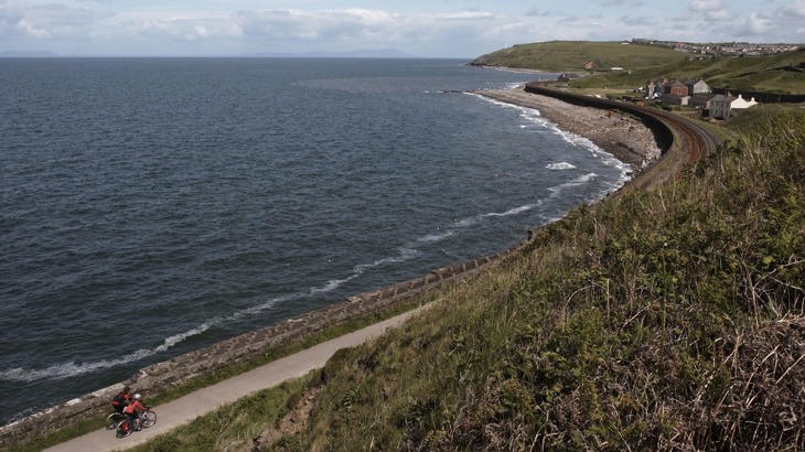 Two cyclists on coastal cycle path with sea wall and beach and buildings in the distance