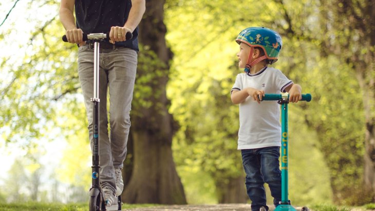 Boy in t shirt, jeans and helmet riding blue scooter with man riding black scooter through park
