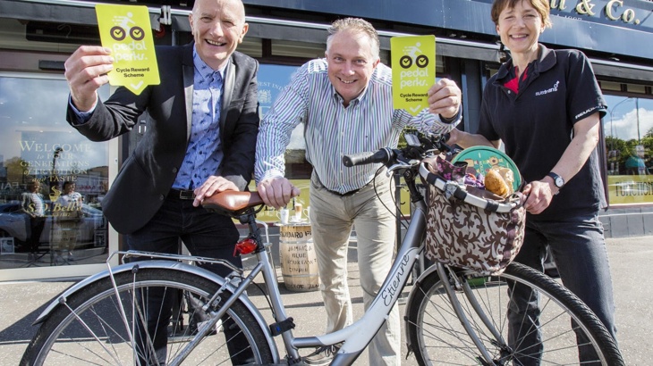 Three people posing with a bike outside a shop with flyers in hand Pedal Perks launch in Northern Ireland