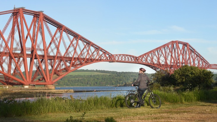 Cyclist in helmet and grey hoodie standing on grassy riverbank, looking at large, red metal bridge