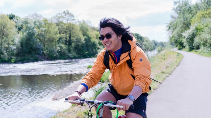 Cyclist in dark glasses and orange jacket on asphalt cycle path by river