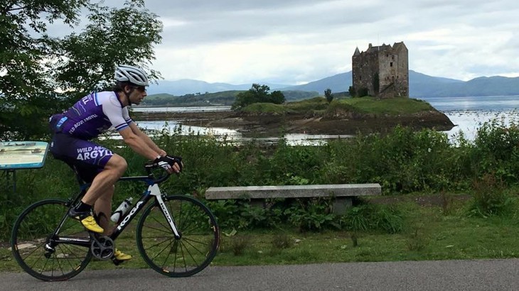 Man cycling on road bike on asphalt path in white helmet and blue and white lycra. To the side is loch with island and castle on it