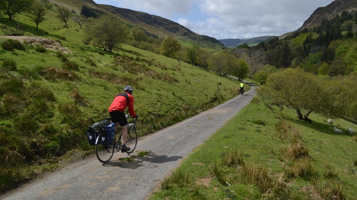 Two road cyclists with panniers, one in red jacket the other in bright green, on gravel track through Welsh mountains