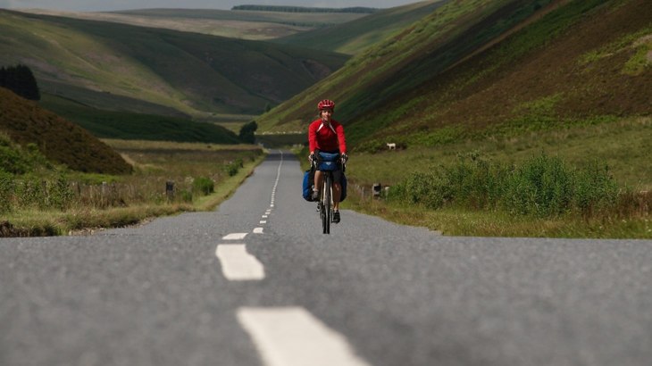 Person riding road bike with panniers wearing red helmet and jersey on road through hills