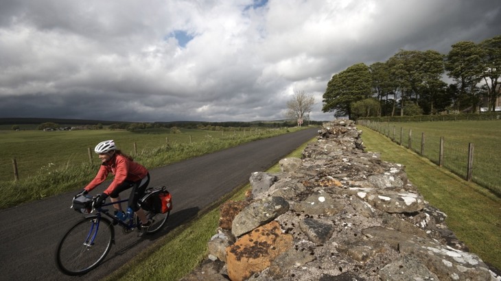 Cyclist on road bike with panniers wearing red jacket and white helmet cycling on road past Hadrian's Wall