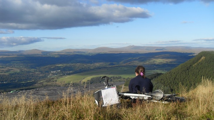 Someone sitting on grassy hillside with bike behind them, looking out on Welsh valley with quarry below