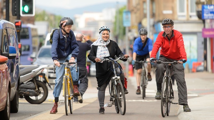 Three adults wearing helmets and riding bikes are waiting at a traffic light in a cycle lane in a city, there are queuing cars beside them and everyone is smiling.