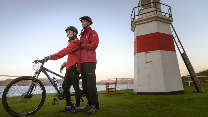 Two people in red coats and cycling helmets, one with mountain bike, standing in front of white and red lighthouse