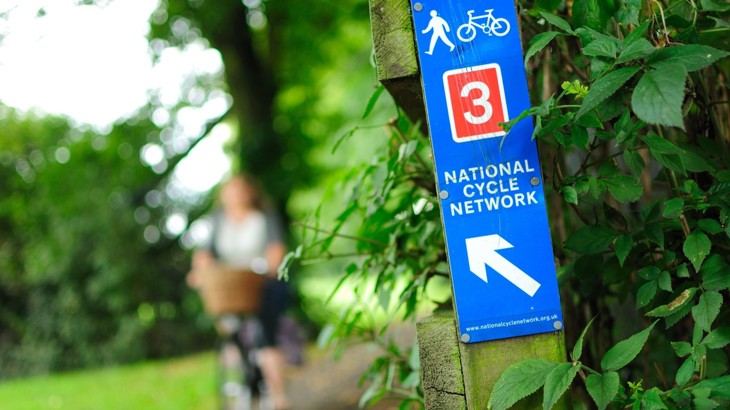 Blue and red sign for National Cycle Network Route 3 with cyclist in background