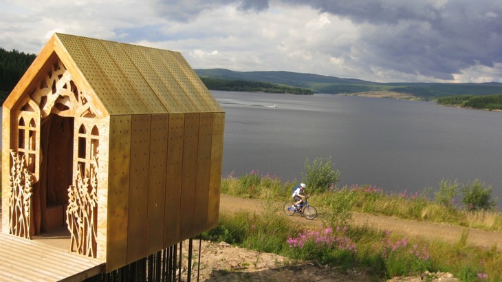 Wooden shed with carvings on the front overlooking cycle path with cyclist on it and lake