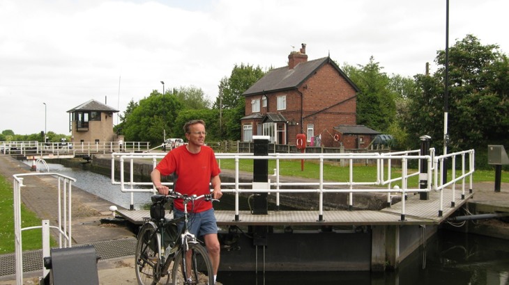Man in t shirt and shorts pushing bike at side of canal locks with red brick house in the background