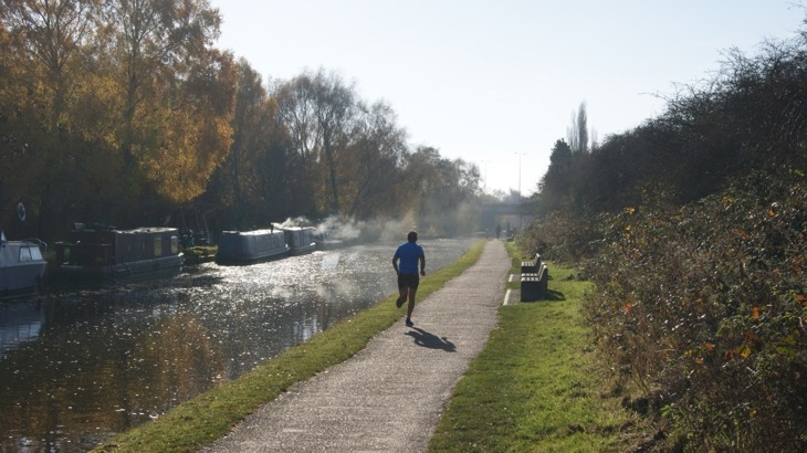 Runner in shorts on canal towpath with narrowboats moored on the opposite bank 