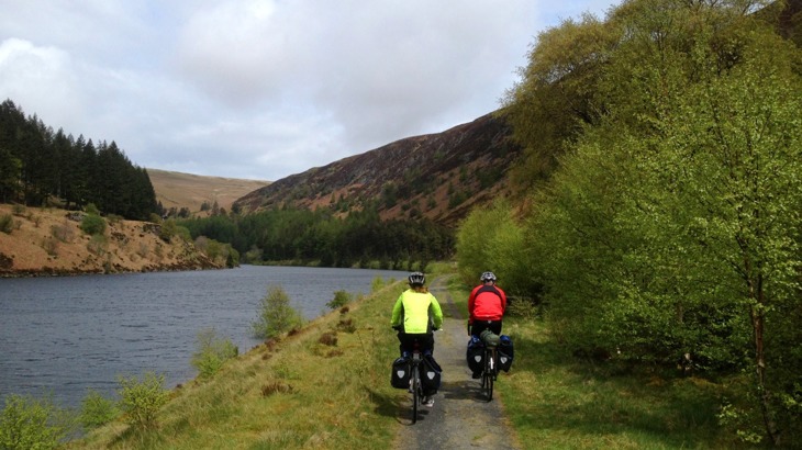 Two cyclists with touring gear on cycle path by river in valley with trees