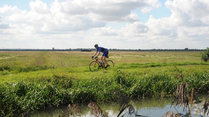 Road cyclist in blue great cycling along path by canal with flat green fields behind 