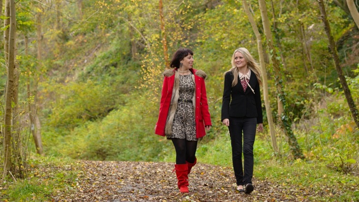 Two women walking on path strewn with fallen leaves through woods