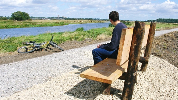 Person sitting on wooden bench by gravel path overlooking river