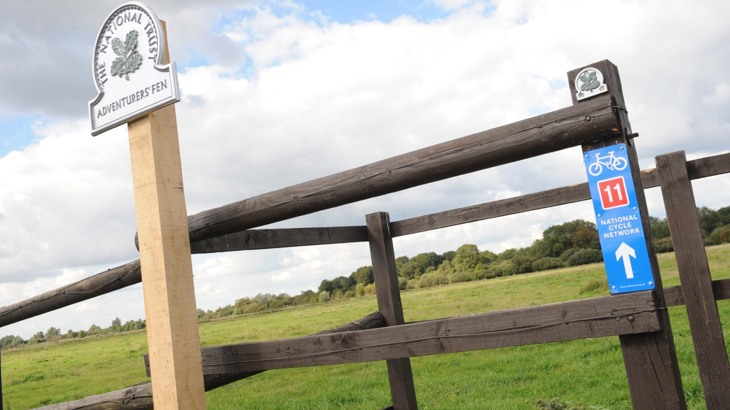 Fence with National Trust sign reading "Adventurers' Fen" and National Cycle Network Route 11 sign