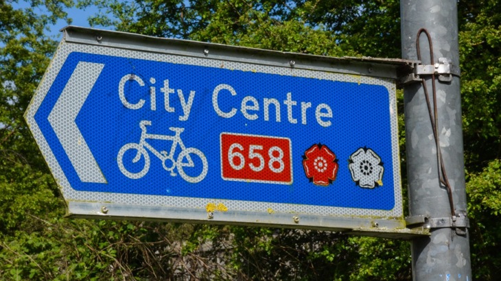 Blue street sign reading "City Centre" with white bicycle symbol, red and white rose symbols, and red box reading "658"
