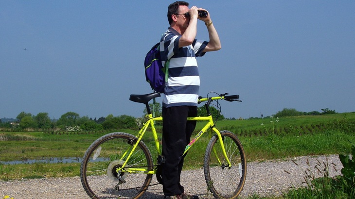 Man with rucksack and bike looking through binoculars in wetland area
