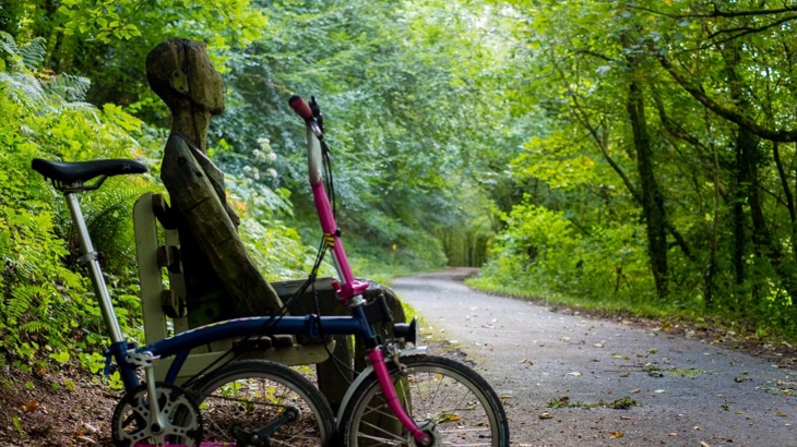 Fold-up bike leaning against bench with wooden sculpture in shape of person, by asphalt path in woodland