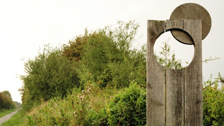 Flat wooden sculpture with circular hole in front of bushes, next to cycle path