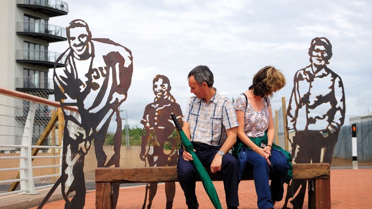 Couple sitting on bench in front of metal artworks shaped like people's silhouettes 