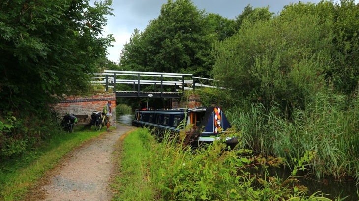 Man standing with bike on canal towpath with narrow boat passing under bridge